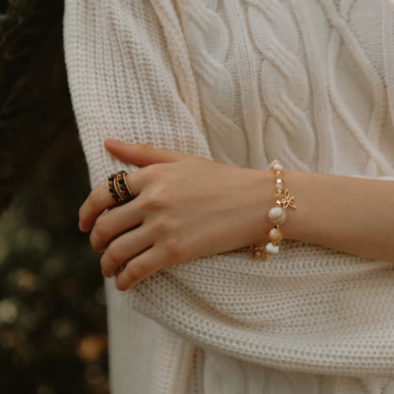 Model with crossed arms wearing the Trochus Shell Bracelet with Lotus from the Flow Collection, showcasing lustrous cream beads for wisdom and wealth against a soft white cable-knit sweater.